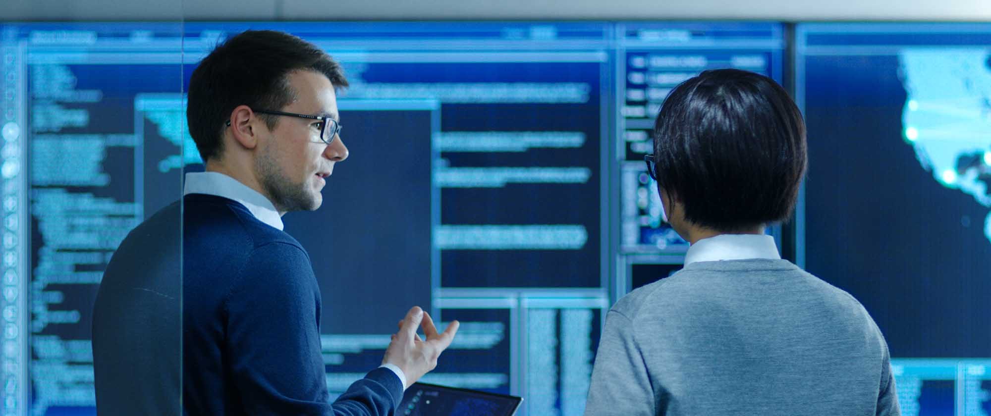 In the System Control Room IT Specialist and Project Engineer Have Discussion while Holding Laptop, they're surrounded by Multiple Monitors with Graphics. They Work in a Data Center on Data Mining.